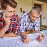 Deux professionnels du bâtiment, un jeune homme au téléphone et un homme mûr tenant une tablette, examinent des plans de construction sur un chantier. Faire appel à un architecte pour rénover sa maison garantit un suivi précis des travaux.