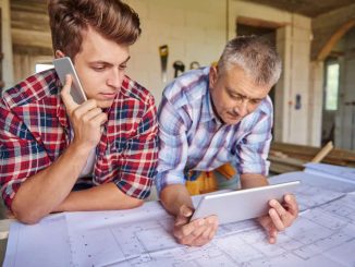 Deux professionnels du bâtiment, un jeune homme au téléphone et un homme mûr tenant une tablette, examinent des plans de construction sur un chantier. Faire appel à un architecte pour rénover sa maison garantit un suivi précis des travaux.