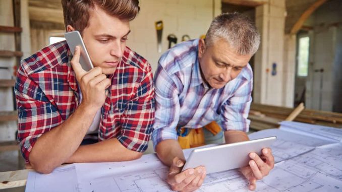 Deux professionnels du bâtiment, un jeune homme au téléphone et un homme mûr tenant une tablette, examinent des plans de construction sur un chantier. Faire appel à un architecte pour rénover sa maison garantit un suivi précis des travaux.