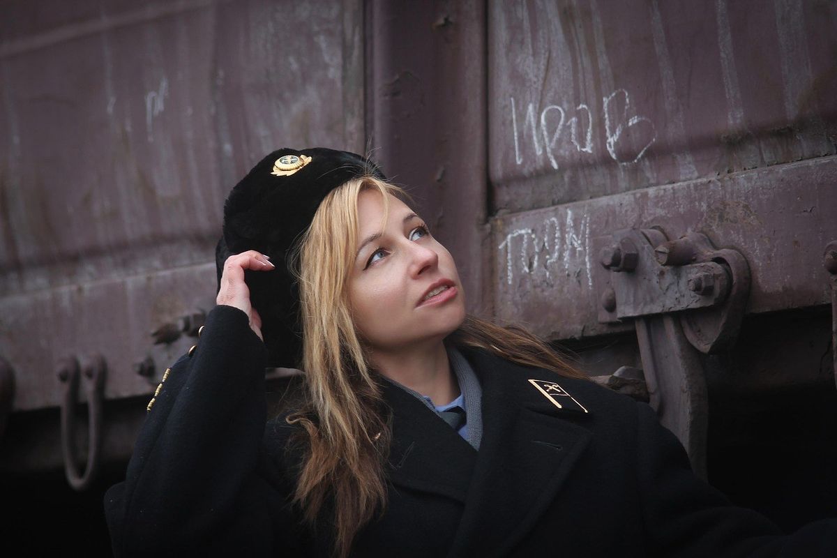 Femme en manteau noir et chapeau inspectant une structure métallique rouillée