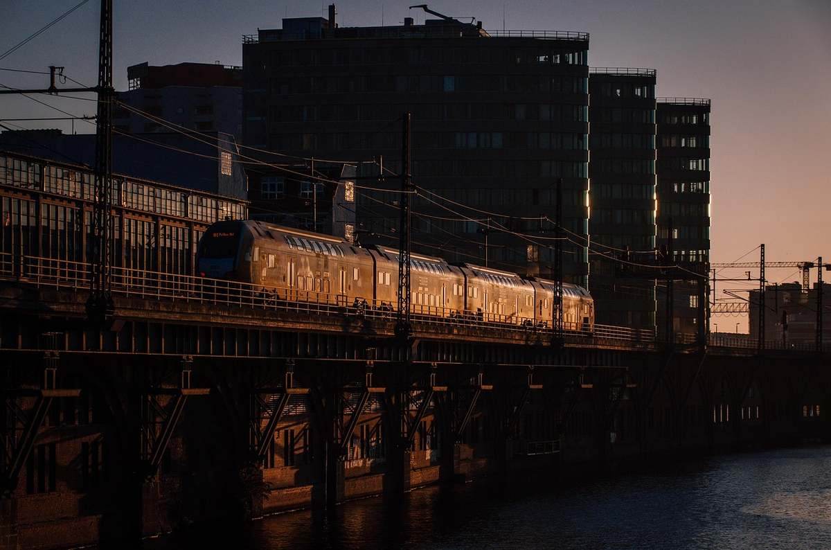 Train de voyageurs illuminé au coucher du soleil sur un pont, avec immeubles urbains en arrière-plan